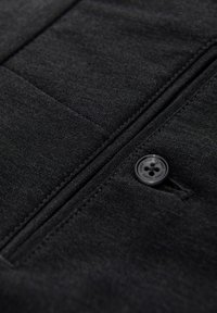 Zwart stoffen detail met een geweven textuur, een verticale rits en een ronde zwarte knop met vier gaten.