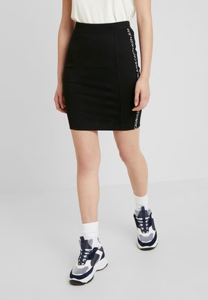 Kobieta ubrana w dopasowaną czarną mini spódniczkę z napisem „Calvin Klein Jeans” po boku, białe skarpetki oraz granatowo-białe trampki, stoi na szarej podłodze.