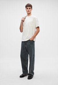 Λευκό κοντομάνικο μπλουζάκι με τσέπη στο στήθος, συνδυασμένο με φαρδιά τζιν παντελόνια σε σκούρο χρώμα και μαύρα παπούτσια, με φόντο ουδέτερου χρώματος.