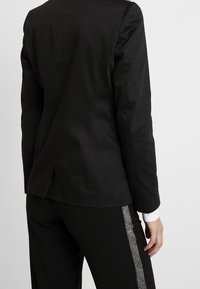 Blazer negro ajustado con mangas largas, que cuenta con una apertura en la parte trasera. Combinado con pantalones negros adornados con una franja plateada a lo largo del costado.