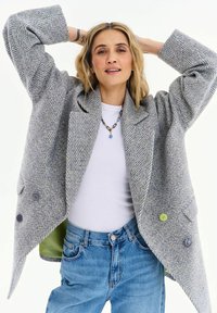 Žena s blond vlasmi v sivom kabáte s rybím vzorom, bielom tričku, modrých džínsoch a náhrdelníku pózujúca s rukami za hlavou.