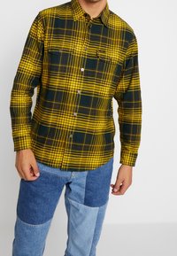 Geel en donkergroen geblokt flanellen overhemd met knoopsluiting, twee borstzakken en een afgeronde zoom, gecombineerd met patchwork spijkerbroek.