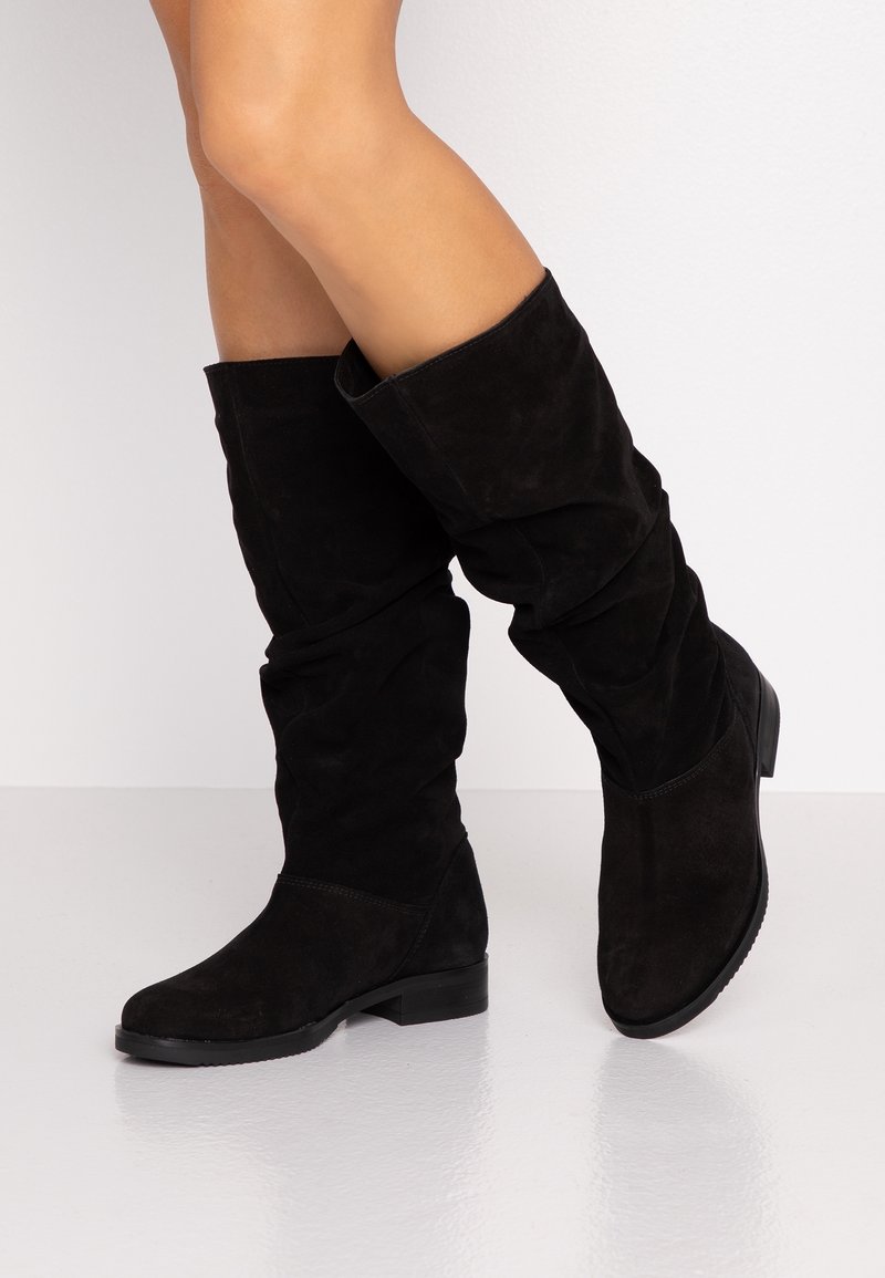 Czarne zamszowe kozaki do kolan noszone na skrzyżowanych nogach, z niskim obcasem i lekko opadającym krojem na jednolitym białym tle.