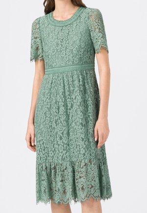 Kvinde iført en mintgrøn knælængde blondekjole med korte ærmer, en figursyet talje og bølgede kanter på skørtekant og ærmer.