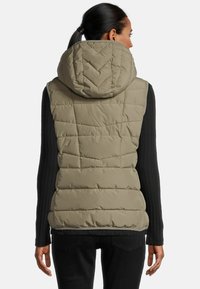 Gilet en duvet khaki avec capuche, présentant un design matelassé et un col montant, associé à un haut noir à manches longues côtelées et à un jean noir.