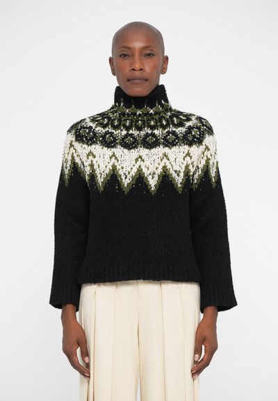 Kvinde iført en sort rullekravesweater med hvide og grønne geometriske mønstre, parret med højtaljede, cremede, plisserede bukser.