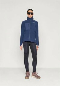 Γυναίκα με ναυτική ζακέτα με φερμουάρ, μαύρα κολάν με λεπτομέρειες, καφέ αθλητικά παπούτσια και μεγάλα μαύρα γυαλιά ηλίου, που στέκεται σε απλό φόντο.