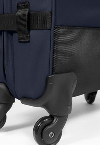 Laavakankainen laukkukassi, jossa on teksturoitu musta pohja, mustat pyörät ja tukeva musta olkahihna. Lähikuva korostaa materiaali- ja muotokäytäntöjä.