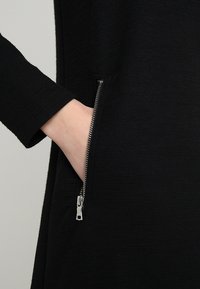 Czarna dzianinowa sukienka z boczną kieszenią i srebrnym zamkiem błyskawicznym. Tekstura wydaje się gładka i subtelna, podkreślając elegancki, minimalistyczny styl.