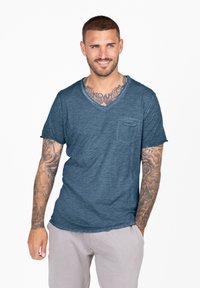 T-shirt blu a maniche corte con scollo a V, tessuto strutturato, dotato di una piccola tasca sul petto, vestibilità rilassata e orlo leggermente curvato.