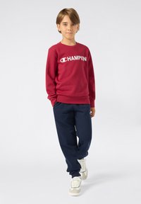 Sudadera roja con logo blanco de "Champion", combinada con pantalones de chándal azul marino y zapatillas beige. Textil suave, corte relajado.