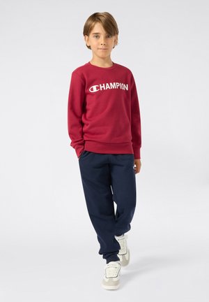 Czerwona bluza z białym logo "Champion", w połączeniu z granatowymi spodniami dresowymi i beżowymi sneakersami. Gładki materiał, luźny krój.