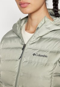 Lys grønne pufferjakke med vandret syninger, lynlås-lukning, hætte og Columbia-logo på venstre side. Glat tekstur.