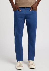 Pantaloni in cotone blu con design a gamba dritta, tasche frontali e una texture liscia, abbinati a sneakers di colore chiaro.