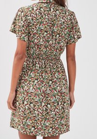 Femme portant une robe à manches courtes à fleurs multicolores avec une taille élastique, se tenant contre un fond clair uni, dos à l'appareil photo.