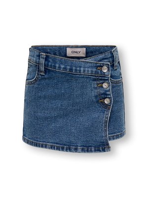 Fille souriante aux cheveux bouclés porte un haut blanc sans manches, une jupe en jean, des sandales marron et tient une veste bleu clair sur une épaule.
