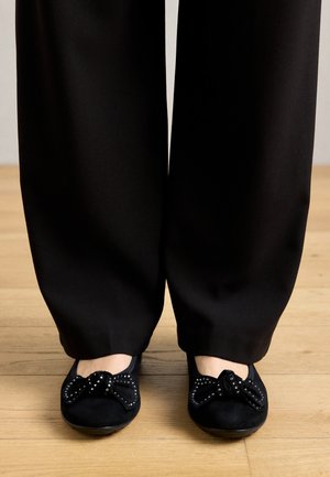 Schwarze Wildleder-Ballerinas mit einer Schleifenakzentuierung, die mit Nieten verziert sind, kombiniert mit weiten schwarzen Hosen. H�ölzerner Boden im Hintergrund.