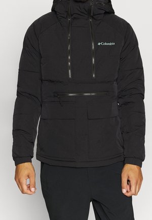 Veste isolante noire Columbia avec capuche, poche poitrine zippée à l'avant et poches kangourou portée par une personne avec les mains visibles.