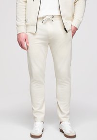 Homme portant un pantalon de jogging couleur crème avec taille à cordon, une veste zippée assortie et des baskets blanches sur un fond uni.