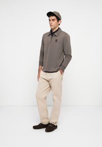 Mann iført beige bukser, brune loafers, grå langermet genser med kvart glidelås og en rutete caps, som står mot en enkel hvit bakgrunn.