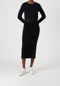 Zwarte langemouwen top gecombineerd met een knielange zwarte rok met een logo tailleband en een zijrits detail, gedragen met witte sneakers.