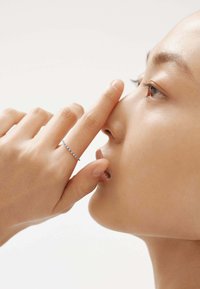 Anillo delgado de plata con pequeñas piedras preciosas, usado en el dedo, cerca de la nariz y la boca de la persona. La piel de la persona es suave.