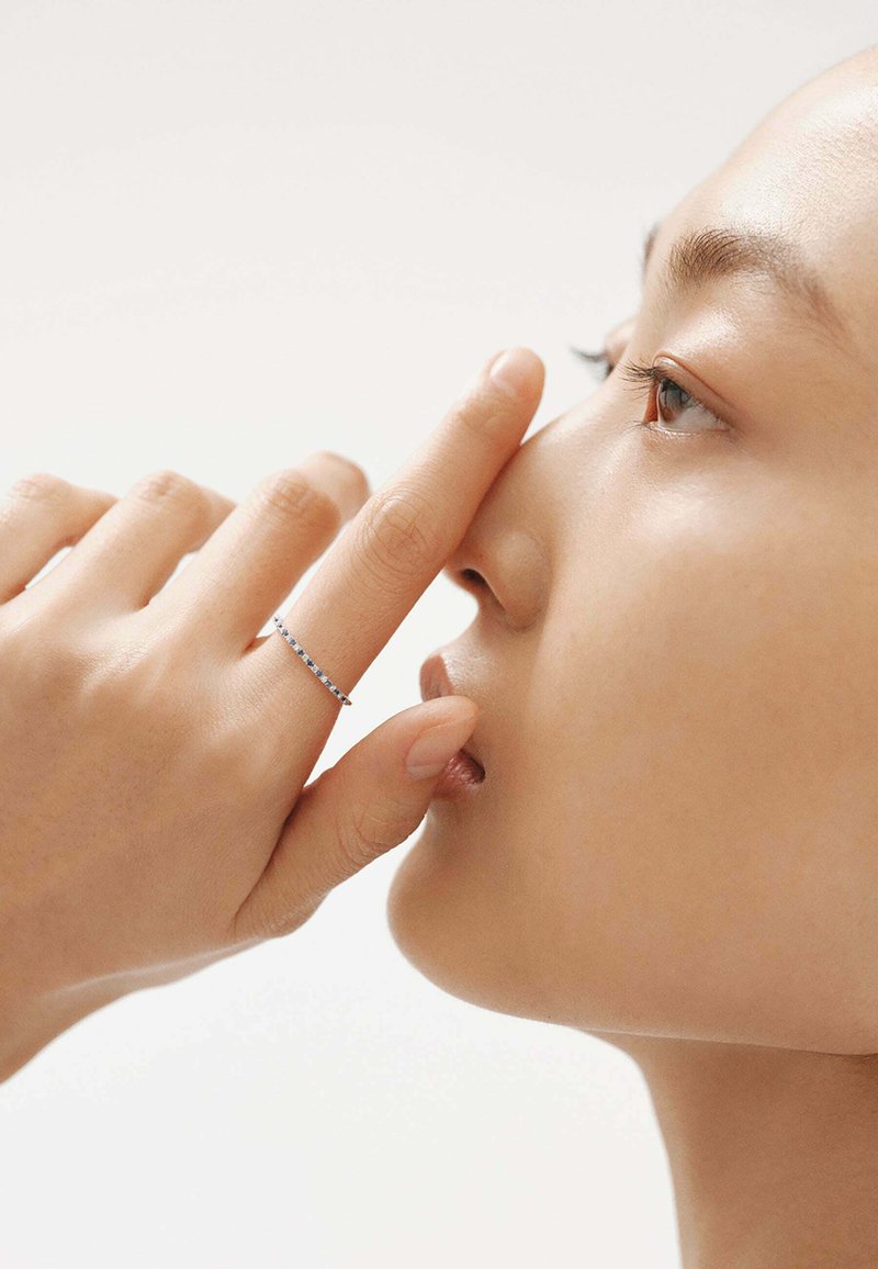 Anillo delgado de plata con pequeñas piedras preciosas, usado en el dedo, cerca de la nariz y la boca de la persona. La piel de la persona es suave.