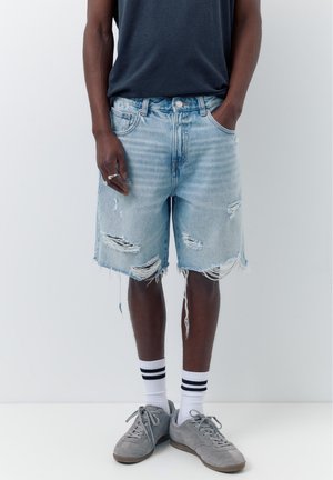 Person trägt hellblaue, zerrissene Jeansshorts, schwarzes T-Shirt, weiße Socken mit schwarzen Streifen und graue Sneakers, steht vor weißem Hintergrund.