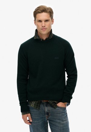 Donkergroene gebreide trui met geribde manchetten, gecombineerd met een zwart-groene geruite shirt. Model draagt spijkerbroek en een horloge.
