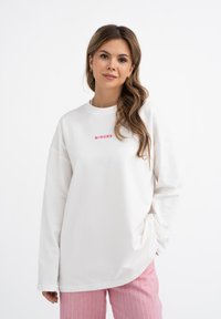 Witte lange mouwen shirt met roze tekst "MIRONS" op de borst. De stof lijkt zacht; heeft een relaxte pasvorm en een ronde hals.