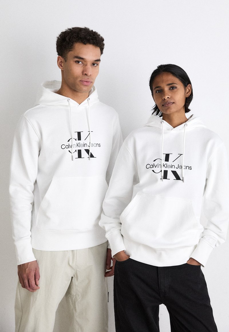 Bijele dukserice s centralnim crnim logom "Calvin Klein Jeans", koje nose dva modela; jedan ima prednju džepicu, mekanu teksturu i opušteni kroj.