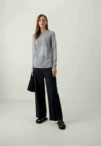 Nainen, jolla on päällään pitkähihainen harmaa paita, mustat leveät housut, mustat sandaalit, ja hän pitää kädessään mustaa tekstiilitaskua, seisoo tasaisella taustalla.