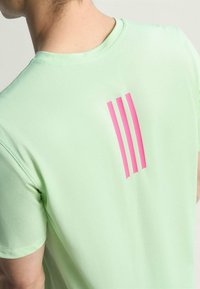 Camiseta deportiva de color verde claro con tres franjas rosas como elemento de diseño en la parte posterior. Fabricada con una tela texturizada y transpirable.