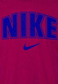 Červené bavlněné tričko s výrazným modrým nápisem "NIKE" a modrým logem swoosh. Hladký povrch bez dalších vzorů.