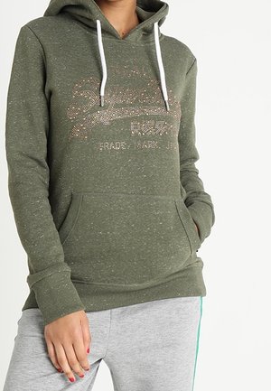 Persoon draagt een olijfgroene hoodie met een glinsterend "Superdry" logo en witte trekkoorden, gecombineerd met lichtgrijze sweatpants met een groene streep.