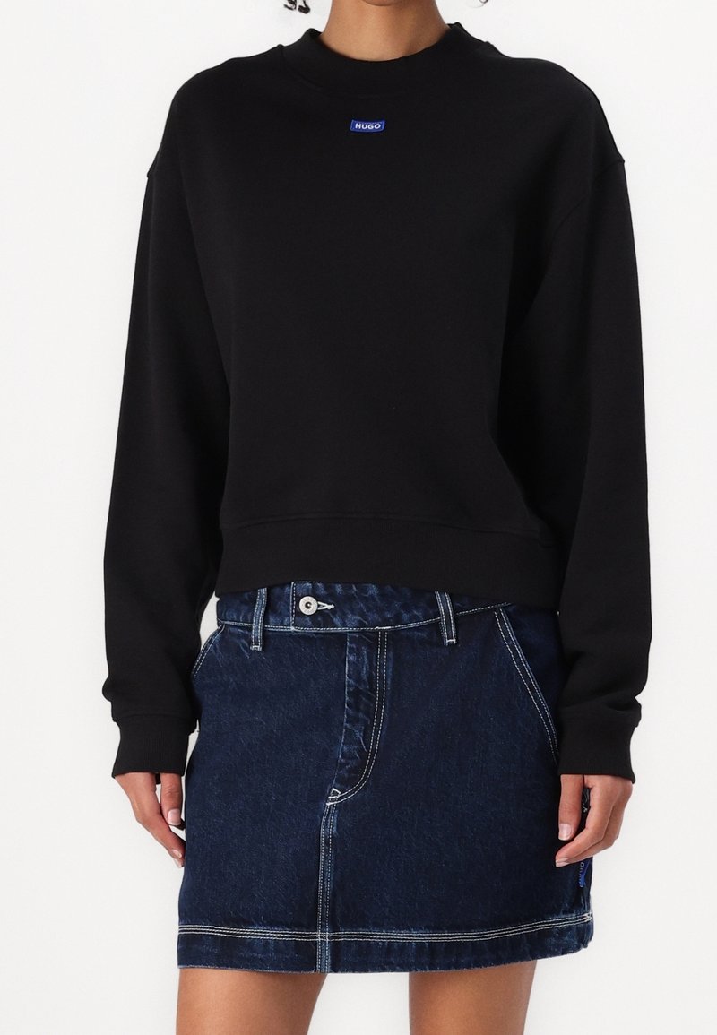 Personne portant un sweat-shirt noir à manches longues avec un petit logo bleu "Hugo" et une jupe en jean bleu foncé avec des coutures visibles.