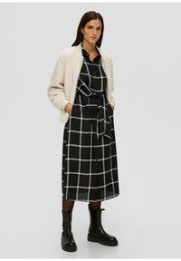 Frau in einem schwarz-weißen karierter, mit einem Gürtel versehenen Kleid, cremefarbener strukturierten Bomberjacke, schwarzen Strumpfhosen und schwarzen klobigen Stiefeln, steht mit den Händen in den Taschen.