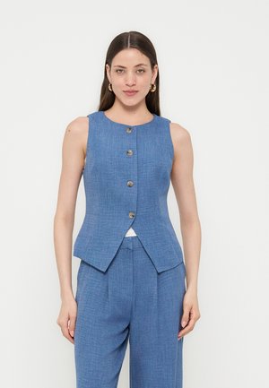 Frau trägt ärmellose blaue Weste mit Knöpfen und passende blaue hoch taillierte Hose, steht vor weißem Hintergrund.