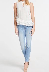 Camiseta sin mangas blanca con rayas verticales y detalle de encaje, combinada con jeans ajustados de color azul claro y tacones nude. Detalle de escote en forma de llave.