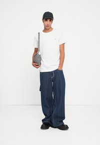 Tânăr îmbrăcat cu un tricou alb, pantaloni largi cu dungi bleu marin, o șapcă neagră și pantofi negri, ținând în mână o geantă gri tip crossbody.