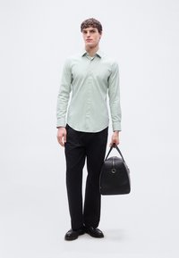 Άνδρας με ανοιχτό πράσινο πουκάμισο με κουμπιά και μαύρο παντελόνι, κρατώντας μια μαύρη δερμάτινη τσάντα duffel στο δεξί χέρι, στέκεται σε απλό λευκό φόντο.