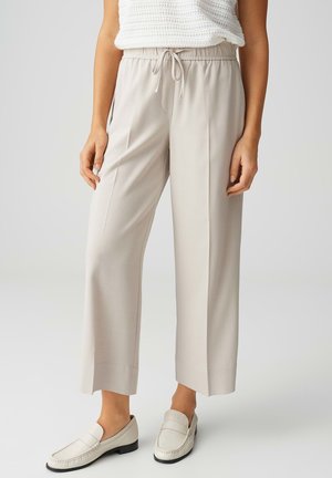 Pantalon large beige court à taille élastique avec cordon, associé à des mocassins blancs, porté par une personne debout les mains détendues.