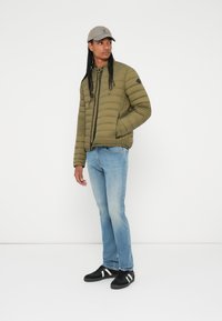 Chaqueta acolchada verde oliva con cierre de cremallera y bolsillos laterales, llevada sobre unos jeans azul claro y zapatillas negras con rayas blancas.