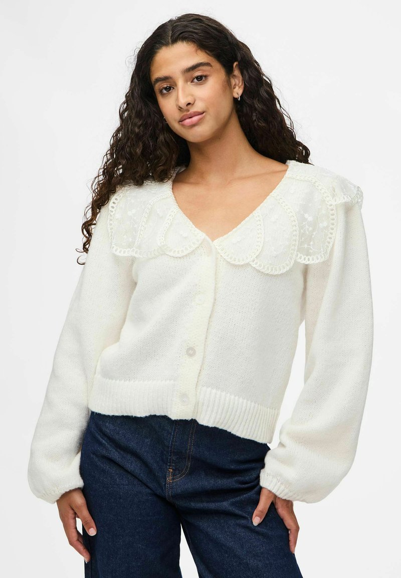 Vit cardigan med en scalloped spetskrage, stora knappar och ribbade manschetter. Mjuk textur med avslappnad passform och långa ärmar.