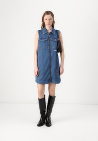 Denim bez piedurknēm kleita ar centrālo rāvējslēdzēju, divām krūšu kabatām un divām sānu kabatām. Valkāta ar melniem spīdīgiem augstiem zābakiem.