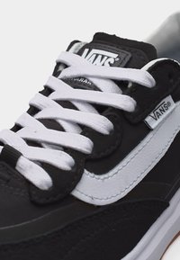 Ασπρόμαυρο αθλητικό παπούτσι Vans με άσπρα κορδόνια, πλαϊνή ρίγα, ετικέτα με το λογότυπο Vans και επενδυμένη γλώσσα, πάνω σε φωτεινό φόντο.