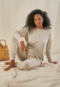 Beige Strickpullover und passende Hose aus weichem Material. Der Pullover hat einen runden Halsausschnitt und gerippte Bündchen. In der Nähe steht ein geflochtener Korb.