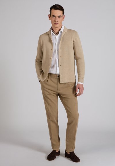 Man draagt een beige vest, wit overhemd, geruite sjaal, zandkleurige broek en donkerbruine loafers, staat met één hand in de zak.
