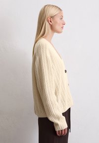 Frau mit glattem blonden Haar, die eine kremfarbene Strickjacke mit schwarzen Knöpfen und dunklen Hosen trägt, im Profil vor einer schlichten Wand stehend.