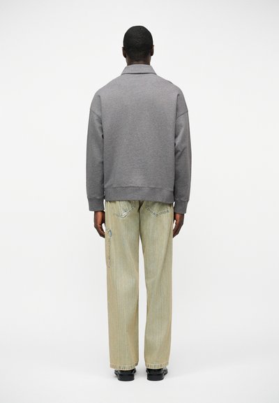 Άνδρας που στέκεται με την πλάτη στραμμένη, φοράει γκρι μπλούζα με μακριά μανίκια και ανοιχτό μπεζ φαρδιά τζιν με μαύρα παπούτσια, σε λευκό φόντο.